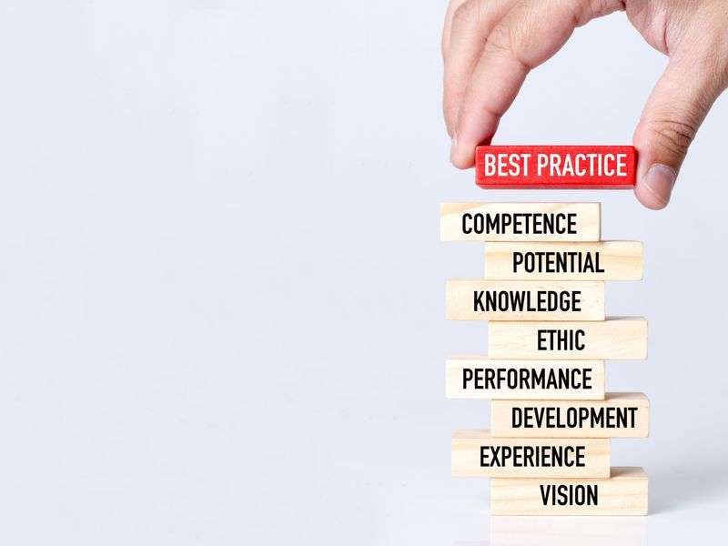 Hand placing a "Best Practice" wooden block atop blocks reading competence, potential, knowledge, ethic, performance, development, experience, and vision