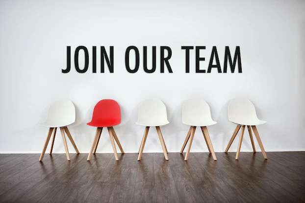 "Join Our Team" sign above a row of white chairs with one red chair, symbolizing recruiting construction talent
