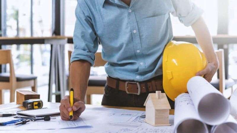 Contractor in a blue shirt writing on blueprints at a desk with a yellow hard hat and model house