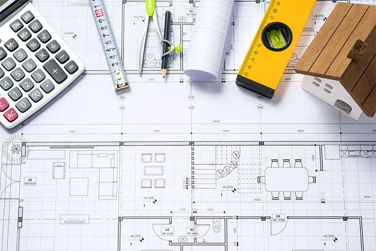 Architectural blueprints with a calculator, ruler, level, and model house on a desk