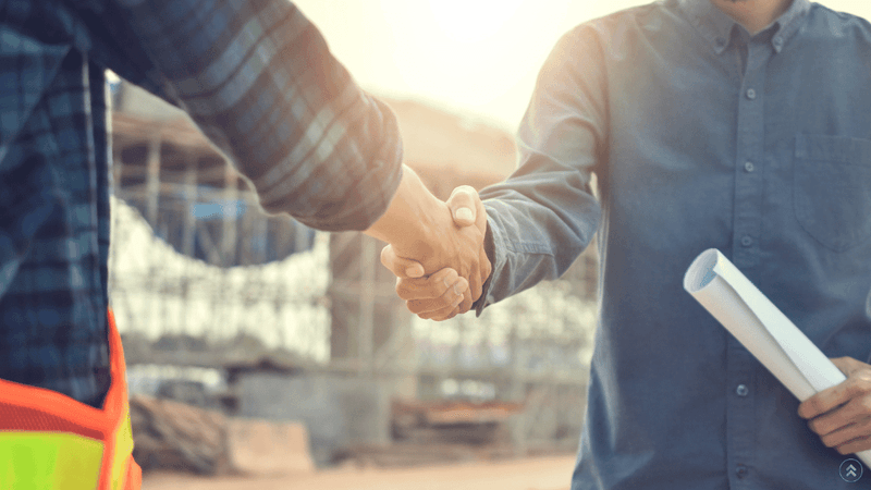 Two construction workers shaking hands on a job site while one holds rolled blueprints