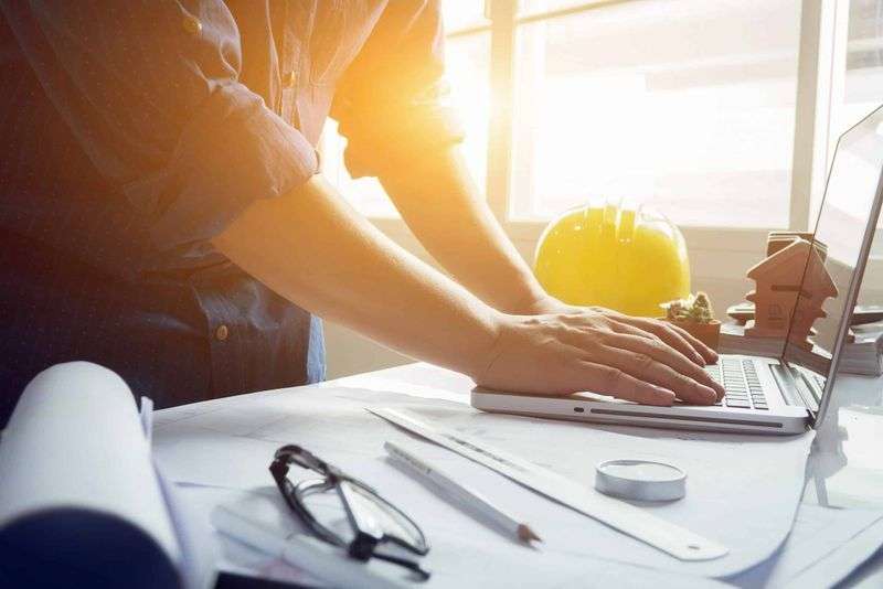 Hand typing on a laptop beside a yellow hard hat on a sunlit desk
