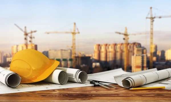 Yellow hard hat with rolled blueprints on a table, with construction cranes and city skyline in the background